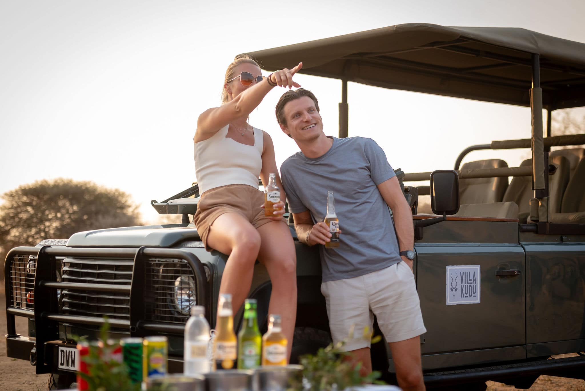 A couple is sitting and standing on the hood of a Land Rover defender in the African wilderness, drinking beer and pointing into the distance. The vehicle has a "Villa Kudu" sign.