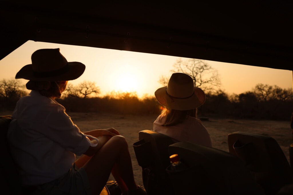 Two people wearing safari hats watch a dramatic, orange sunset over the dry, tree-dotted African landscape from the seats of an open safari vehicle.