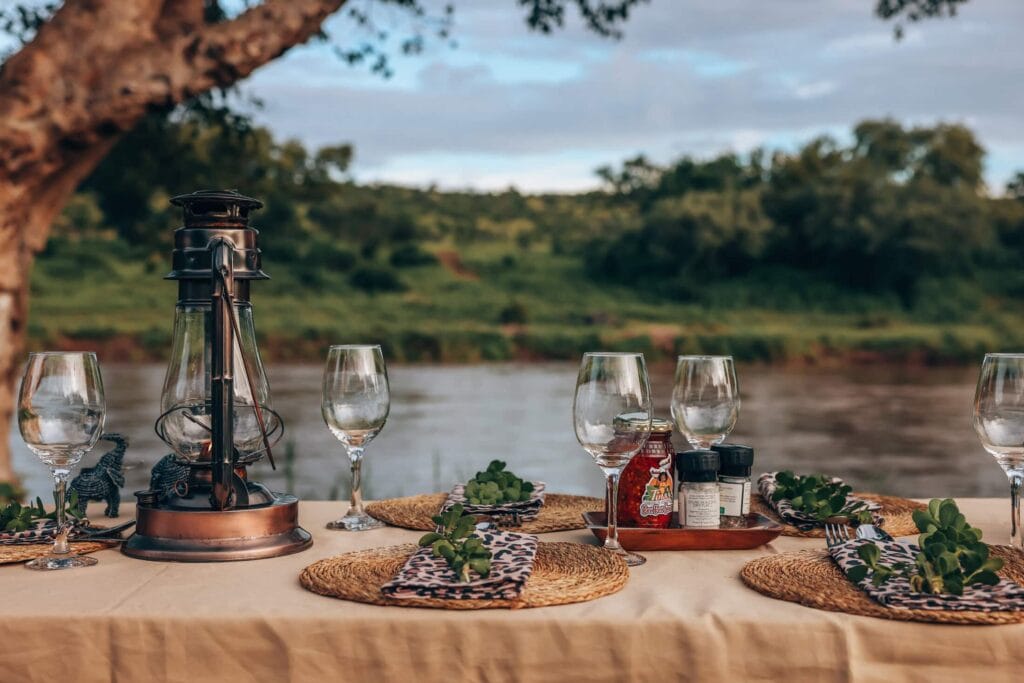 Detailed view of a luxury safari dinner table setting by a river, featuring woven placemats, animal-print napkins, wine glasses, and a copper lantern.