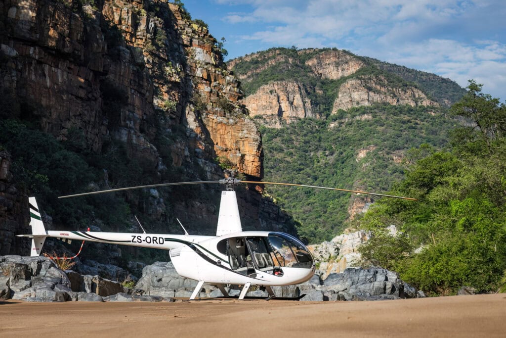 A white helicopter with side doors open and registration ZS-ORD on the tail boom is parked on a sandy riverbank in a deep gorge, with towering, layered rock cliffs and green trees in the background.